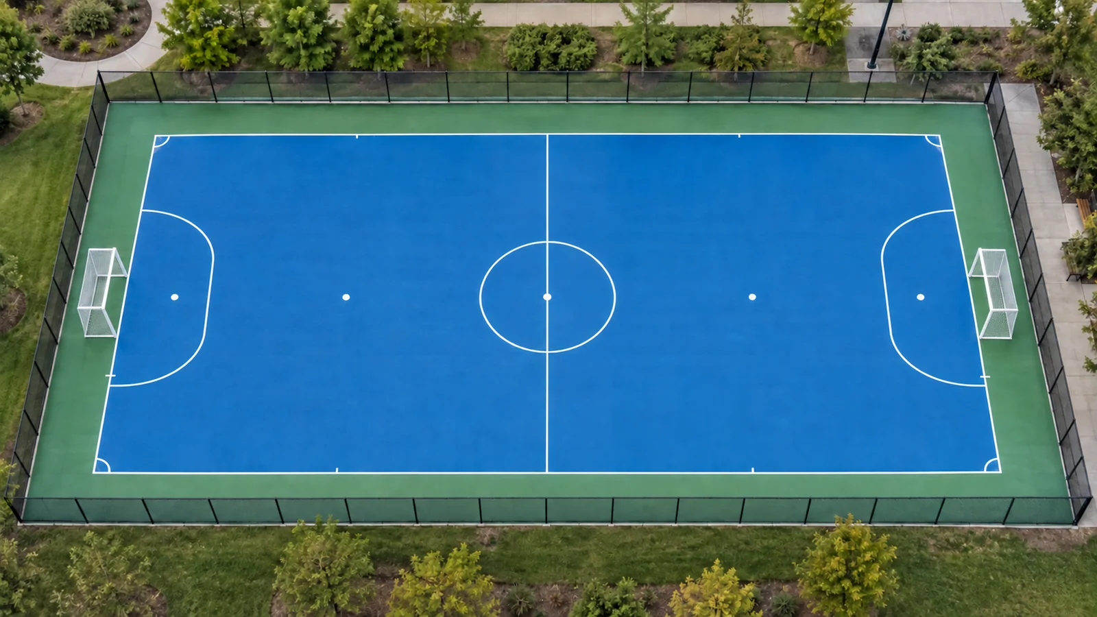 Outdoor blue futsal court with white markings, goals, green apron, fencing, and lighting